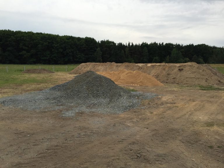 Schotter, Sand- und Erdhügel liegen auf einer Wieser und im Hintergrund ein Wald.